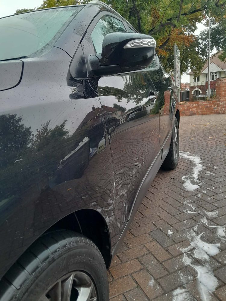 The side of a dark car being washed on a driveway, with foam running off the paintwork